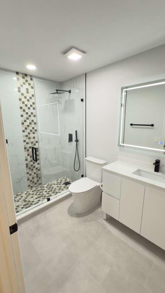 Baño con mampara de vidrio, revestimiento estampado, tocador blanco y piso gris